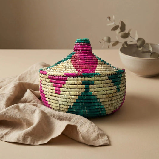 A colorful teal and fuchsia hand-woven Mazari palm basket with a conical lid, styled on a tan linen cloth.